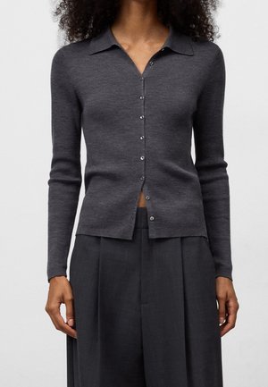 Femme portant un cardigan ajusté à manches longues en charbon avec des boutons et un col, ainsi qu'un pantalon large et taille haute de couleur foncée.