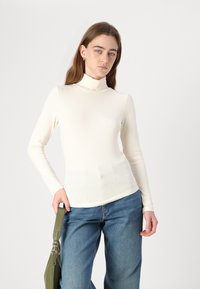 Kremfarget ribbestrikket turtleneck-genser med lange ermer, kombinert med lyseblå vide jeans. Modellen holder en grønn håndveske.