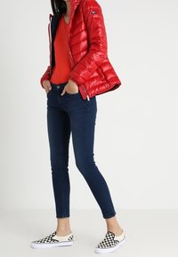 Giacca a vento rossa con finitura lucida, maglietta arancione, jeans skinny blu scuro e scarpe slip-on a scacchi bianco e nero.