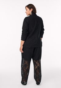 Completo in pizzo nero composto da un blazer su misura e pantaloni a gamba larga con inserti in pizzo. I dettagli in tessuto testurizzato sono molto evidenti.