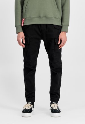 Personne portant un pantalon cargo noir avec plusieurs poches, des poignets élastiques, des baskets noires et blanches, et un sweat-shirt vert olive.