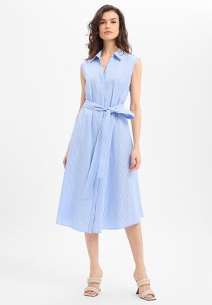 Frau trägt ein ärmelloses, hellblau-weiß gestreiftes Midikleid mit Kragen und gebundenem Taillengürtel, kombiniert mit beigen Sandalen mit Absatz.