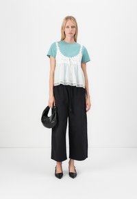 Lichtblauw T-shirt onder een witte top met kant, gecombineerd met zwarte wijde broek en zwarte puntige hakken, terwijl ze een zwarte tas vasthoudt.