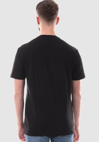 Camiseta de algodón negra con mangas cortas, dobladillo recto y un ajuste clásico, exhibida desde la parte posterior contra un fondo neutro.