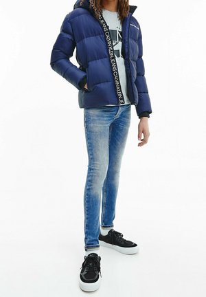 Persona che indossa una giacca a vento blu, jeans azzurri chiari, sneaker bianche e nere e una maglietta grigia Calvin Klein, in piedi contro uno sfondo bianco.