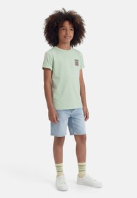 Göndör hajú gyerek világoszöld pólóban, farmer rövidnadrágban, csíkos zokniban és fehér sportcipőben, fehér háttér előtt áll.