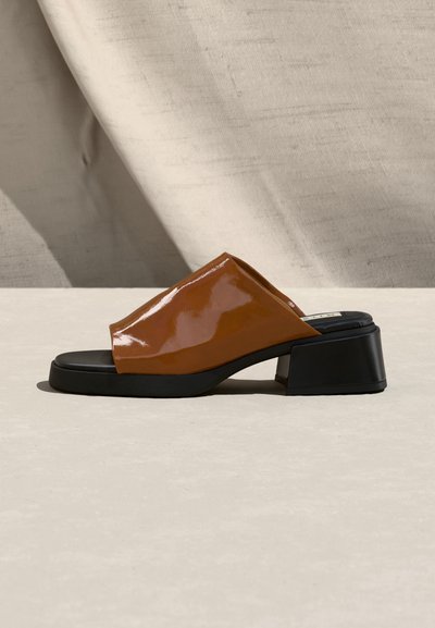 Καφέ λουστρίνι mule με ανοιχτά δάχτυλα, μαύρο χοντρό χαμηλό τακούνι και μαύρη σόλα, τοποθετημένο πλάγια σε φωτεινή επιφάνεια.