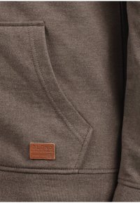Sudadera marrón con un bolsillo frontal, que presenta una superficie texturizada y un dobladillo acanalado. Se adjunta un pequeño parche de cuero etiquetado "BLEND".