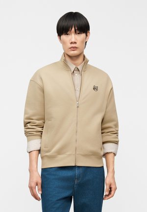 Mladi moški s črnimi lasmi, oblečen v bež jakno na zadrgo z živalsko značko, pod njo bež majico in modre jeans hlače, stoji pred belim ozadjem.