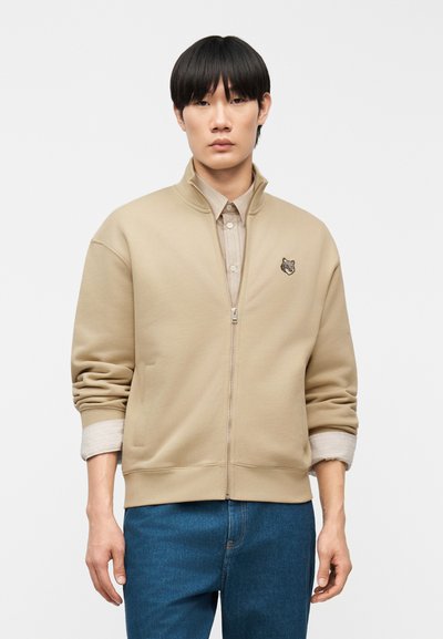 Jeune homme aux cheveux noirs portant une veste beige à fermeture éclair avec un écusson d'animal, une chemise beige en dessous et un jean bleu, debout devant un fond blanc.