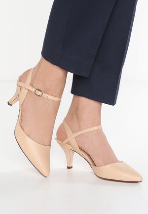 Beżowe buty na niskim obcasie typu kitten heel z paskami wokół kostki i krzyżującymi się, noszone z granatowymi, krótkimi spodniami na jasnym tle.