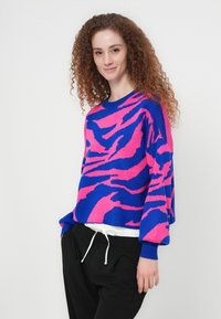 Γυναίκα με σγουρά κόκκινα μαλλιά που φοράει μπλε και ροζ πουλόβερ με σχέδια και μαύρο παντελόνι, στέκεται μπροστά σε απλό λευκό φόντο.