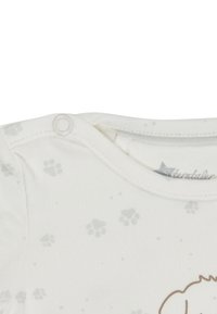 Weißer Baby-Body mit grauen Pfotenabdrücken und einem Druckknopfverschluss an der Schulter, mit einem Teil einer braunen Tierillustration.