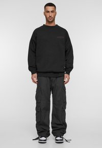 Hanorac negru cu guler rotund și logo violet pe piept. Asortat cu pantaloni cargo cu croi larg, care au multiple buzunare.