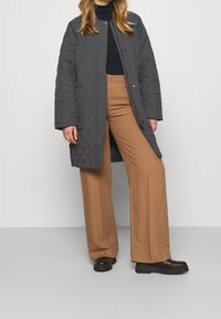 Cappotto lungo grigio trapuntato con chiusure a scatto, abbinato a pantaloni larghi marroni e stivali neri chunky. Tessuto liscio e design a vestibilità ampia.