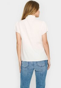 Lichtcrème blouse met korte, kapmouwen en een gerimpelde halslijn. De achterkant laat een losse pasvorm en een zachte stofstructuur zien. Gecombineerd met blauwe jeans.