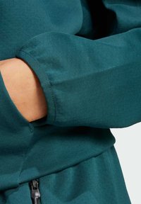 Tyrkysová mikina, mäkká tkanina, detailný záber na manžetu s elastickým zakončením, prešitý lem a bočná vrecko s detailom na zips.