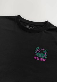 Tricou negru din bumbac cu un grafic colorat al unei pușculițe înconjurată de bani verzi, având text coreean dedesubt, în roz. Design clasic cu gât rotund.