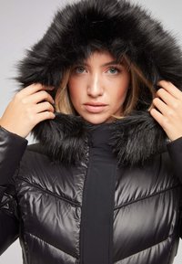 Chaqueta puffer negra con una capucha de pelaje sintético negro y esponjoso, que presenta un diseño acolchado y una textura suave y brillante. Ajuste regulable alrededor de la cara.