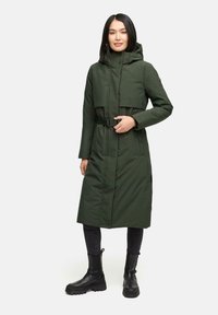 Cappotto imbottito lungo di un verde scuro, con cappuccio e vita cintata. Presenta bottoni a pressione e una silhouette slanciata, indossato sopra pantaloni neri e stivali.