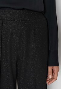 Närbild av svarta byxor med diskreta silvriga glitters detaljer, hög midja, och en svart långärmad topp med manschetter.