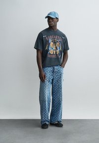 Muž nosiaci tmavé tričko s grafikou, svetlomodré voľné džínsy so vzorom, čierne topánky a svetlomodrú šiltovku stojaci pri obyčajnej stene.