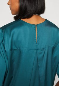 Teal satinbluse med en nøgleåbning i ryggen, afslappet pasform, korte ærmer og diskret tekstur.