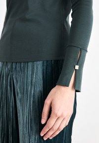 Dark green long-sleeve top features a split cuff with silver hardware detail. Underneath, a deep teal pleated skirt adds texture and contrast.