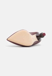 Chaussure à talon haute bordeaux avec un bout pointu, dessus en cuir lisse et semelle nude. Accent rond notable à l'arrière du talon.