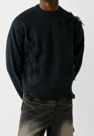 Pull en maille noire avec des déchirures effilochées intentionnelles, porté sur un jean foncé délavé, présenté du cou jusqu'aux hanches sur un fond clair uni.
