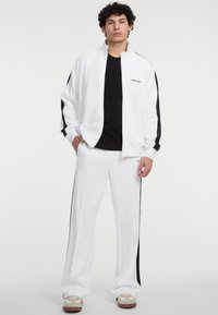 Witte sportoutfit met een ritsjack met zwarte accenten, gecombineerd met bijpassende broek; de stof lijkt glad en licht van gewicht.
