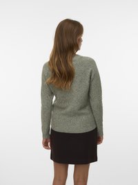 Donna con lunghi capelli rossi, che indossa un maglione grigio a maglia e una gonna nera, in piedi di spalle contro uno sfondo bianco.