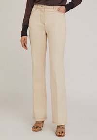 Jean évasé beige avec des coutures visibles, taille haute, cinq poches et une texture lisse, associé à des sandales à talons hauts avec lanières.