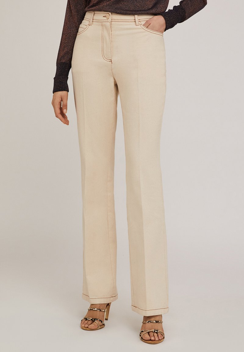 Jean évasé beige avec des coutures visibles, taille haute, cinq poches et une texture lisse, associé à des sandales à talons hauts avec lanières.