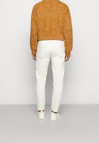 Brązowa koszula w wzory z długim rękawem, białe zwężane dżinsy i białe trampki. Koszula posiada teksturowany wzór; dżinsy są jednolite z tylnymi kieszeniami.