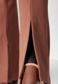 Pantalon marron avec fentes latérales révélant une chaussure à talon bloc en bois avec dessus noir et bordure beige.