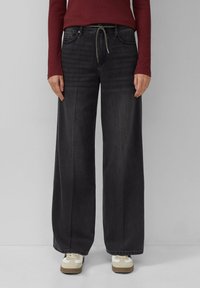 Schwarze weit geschnittene Jeans mit hoher Taille, ausgestattet mit Fronttaschen, einem Kordelzug und einer glatten Textur, kombiniert mit weißen Sneakers.