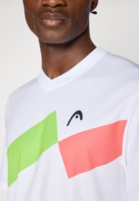 Hvid polo-shirt med grønne og pink geometriske mønstre på fronten. Har en V-udskæring og et lille sort logo på brystet.