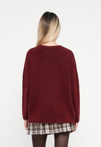 Bordeaux oversized sweater met dropped shoulders, zachte textuur en een ronde halslijn, gecombineerd met een geplooide ruitrok en zwarte legging.
