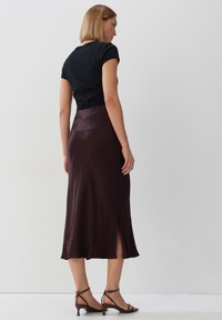 Schwarzes, kurzärmliges Oberteil kombiniert mit einem knielangen, dunklen, satinierten Rock mit seitlichem Schlitz und fließender Silhouette, getragen mit braunen Absatzschuhen.