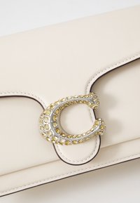 Cremfarbene Leder-Clutch mit einem mit silbernen und gelben Kristallen verzierten Verschluss. Sie verfügt über kontrastierende dunkle Nähte und eine glatte, strukturierte Oberfläche.