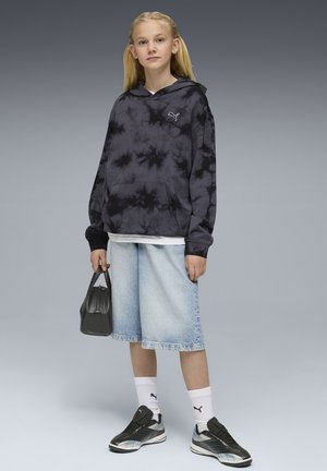 Junge Person trägt einen schwarzen Hoodie mit Batikmuster, helle Jeansshorts, weiße Puma-Socken, schwarze Turnschuhe und hält eine kleine schwarze Tasche.