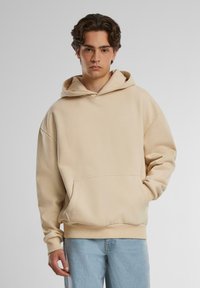 Beige hooded sweatshirt with a front pocket, made of soft fabric, featuring a relaxed fit and ribbed cuffs. Worn with light blue jeans.