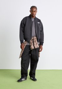 Veste bomber noire avec un logo, t-shirt gris, pantalon baggy noir et baskets noires avec des rayures blanches. Flanelle attachée autour de la taille.
