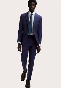Costume deux pièces bleu marine à coupe slim, comprenant une veste à simple boutonnage, des revers crantés et un pantalon à plis plats. Porté avec une chemise blanche et une cravate bleue.
