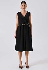 Zwarte mouwloze wikkeljurk met een diepe V-hals, een op de taille cinched stijl met een gouden gesp, en een A-lijn rok. Gedragen met puntige bordeauxkleurige hakken.