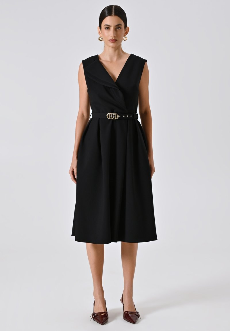 Zwarte mouwloze wikkeljurk met een diepe V-hals, een op de taille cinched stijl met een gouden gesp, en een A-lijn rok. Gedragen met puntige bordeauxkleurige hakken.