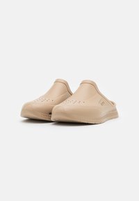 Beige gummiklumpor med stängd tå, slip-on design, ventilationshål på ovansidan och texturerad sula för grepp.