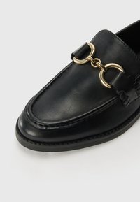 Černé kožené loafers s kulatou špičkou, prošívanými detaily a zlatým řetízkem přes svršek. Hladký povrch s plochou podrážkou.