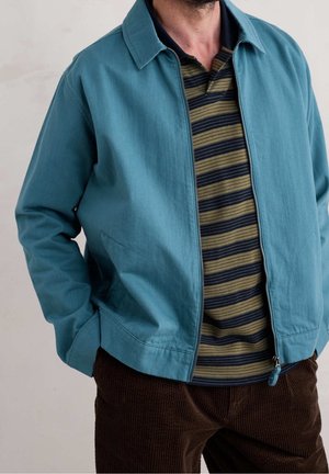 Muž v tyrkysové bundě na zip přes černo-olivově pruhované polo tričko a hnědé manšestrové kalhoty, ruce v kapsách bundy.